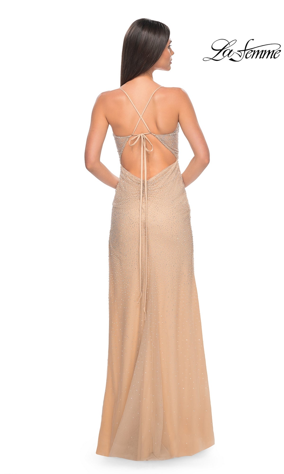 A woman wears the Sheer-Waist Long Beaded Prom Dress (La Femme 32435), a backless, floor-length beige gown with thin crisscross straps and subtle embellishments, standing against a white background.